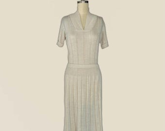May include: A beige knit dress featuring a V-neckline, short sleeves, and a vertical ribbed texture throughout the bodice and skirt. The garment is displayed on a mannequin, showcasing a fitted waist and a flared, knee-length skirt silhouette.