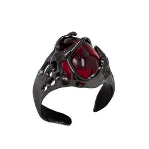 May include: A dark gray ring featuring a large, oval, deep red gemstone. The ring's setting has an intricate, organic design, with a textured, openwork pattern. The band is a simple, smooth curve.