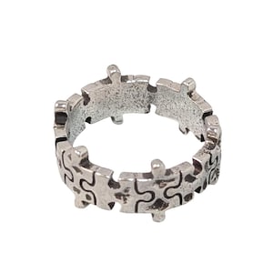 Op de afbeelding: Zilverkleurige ring met een puzzelstukjesontwerp. De ring is gemaakt van metaal met een licht getextureerd oppervlak. De puzzelstukjes zijn in elkaar grijpend en vormen een doorlopende band.