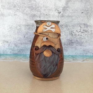 May include: A brown ceramic vase with a pirate face design. The vase features a sculpted pirate hat with a skull and crossbones, a long beard, and a mustache. The vase is approximately 8 inches tall.