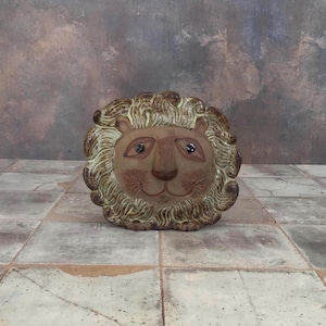 May include: A ceramic lion head sculpture with a detailed mane and facial features. The lion's face is a light brown color, with dark eyes and a smiling expression. The mane is a textured, light brown and green color. The sculpture is on a tiled surface.