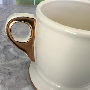 Puede incluir: Taza de cer&aacute;mica color crema con asa y borde dorados. La taza tiene una forma cl&aacute;sica con una base ancha y una parte superior redondeada. El asa es una caracter&iacute;stica distintiva que a&ntilde;ade un toque de elegancia al dise&ntilde;o.