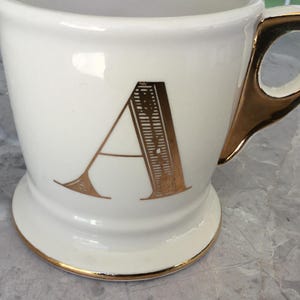 Puede incluir: Taza de cer&aacute;mica blanca con asa y ribetes dorados. La letra "A" dorada se muestra en la parte delantera. La taza tiene una base.