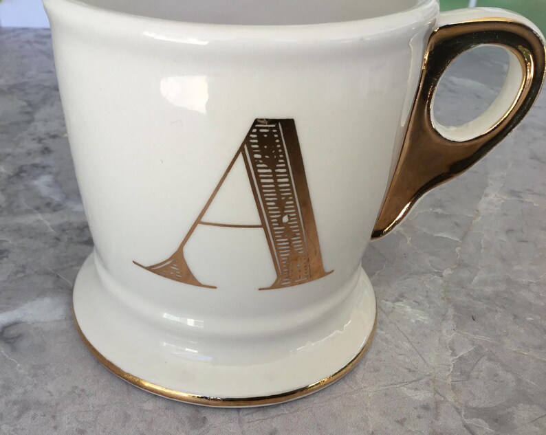 Puede incluir: Taza de cer&aacute;mica blanca con asa y ribetes dorados. La letra "A" dorada se muestra en la parte delantera. La taza tiene una base.