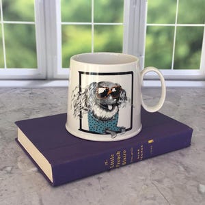 May include: A white ceramic mug featuring a dog wearing sunglasses and a blue polka dot shirt. The mug is set on a purple book titled "The Little French Bistro". The mug has a black border around the dog illustration.