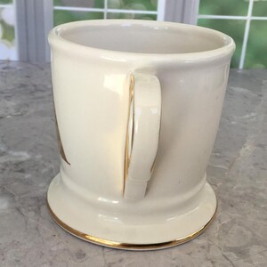 Puede incluir: Taza de cer&aacute;mica color crema con borde y detalles dorados en el asa. La taza tiene una base ancha y un asa curvada. El interior es blanco. La taza es probablemente vintage o antigua.