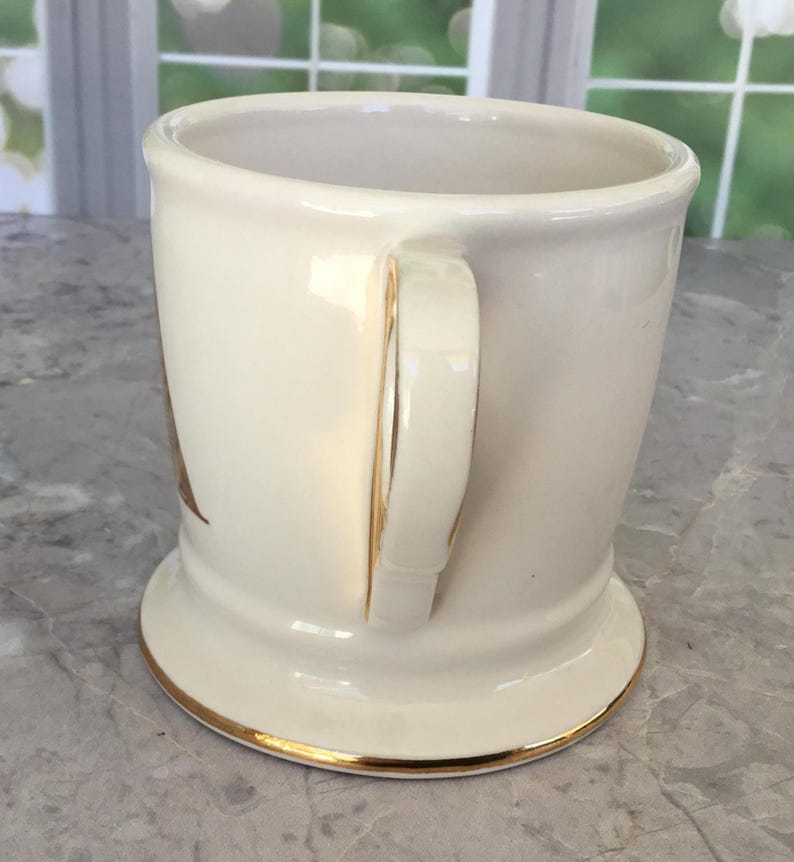 Puede incluir: Taza de cer&aacute;mica color crema con borde y detalles dorados en el asa. La taza tiene una base ancha y un asa curvada. El interior es blanco. La taza es probablemente vintage o antigua.