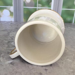 Puede incluir: Taza de cer&aacute;mica color crema con borde y asa dorados. La taza tiene forma cil&iacute;ndrica y un acabado liso y brillante. El asa tambi&eacute;n es de color crema con detalles dorados. La taza est&aacute; vac&iacute;a y parece limpia.