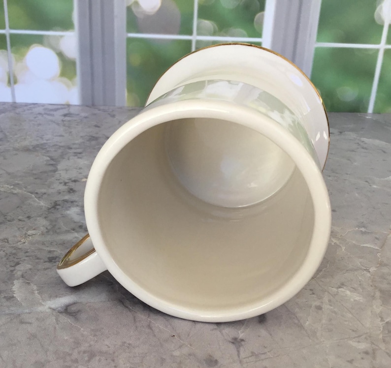 Puede incluir: Taza de cer&aacute;mica color crema con borde y asa dorados. La taza tiene forma cil&iacute;ndrica y un acabado liso y brillante. El asa tambi&eacute;n es de color crema con detalles dorados. La taza est&aacute; vac&iacute;a y parece limpia.