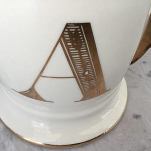 Puede incluir: Taza de cer&aacute;mica blanca con asa y ribetes dorados. La letra "A" se muestra en dorado en la parte delantera. La taza tiene una base de pedestal.