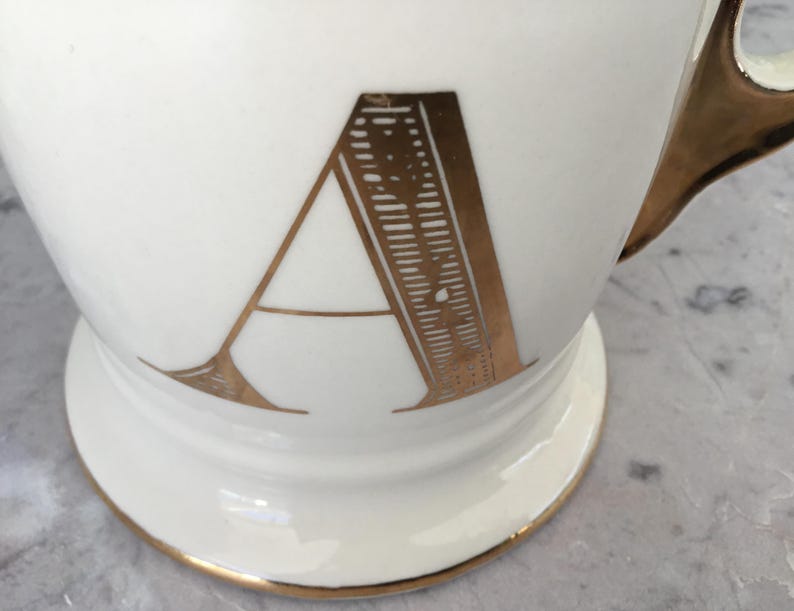 Puede incluir: Taza de cer&aacute;mica blanca con asa y ribetes dorados. La letra "A" se muestra en dorado en la parte delantera. La taza tiene una base de pedestal.