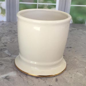 Puede incluir: Un vaso de cer&aacute;mica color crema con un borde dorado en la base. El vaso tiene una forma ligeramente acampanada y un acabado liso y brillante. El interior es de color crema a juego.