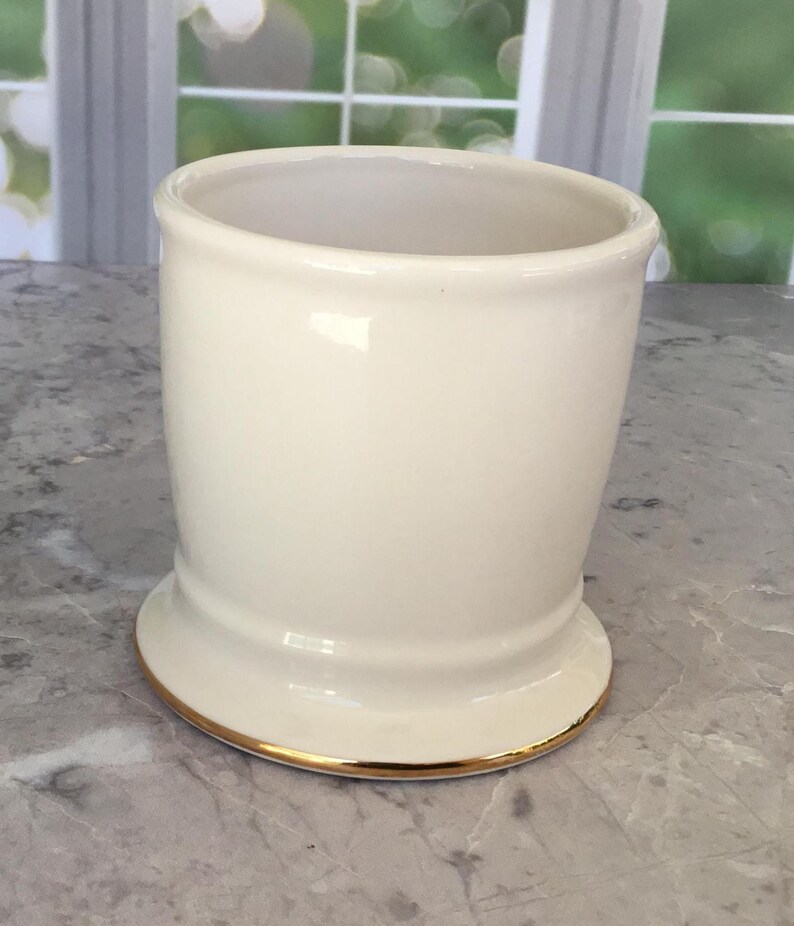 Puede incluir: Un vaso de cer&aacute;mica color crema con un borde dorado en la base. El vaso tiene una forma ligeramente acampanada y un acabado liso y brillante. El interior es de color crema a juego.