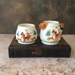 May include: Cream-colored ceramic sugar bowl and creamer set. The creamer has a bear head spout and a handle with a black and white design. Both pieces feature a hunting scene with riders on horseback, dogs, and trees.