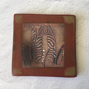 May include: A square, ceramic tile with a raised relief of a stylized cat. The cat has brown and tan stripes and is surrounded by tall grass with white and pink accents. The tile has a reddish-brown border.