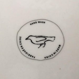 Puede incluir: Superficie de cer&aacute;mica blanca con un dise&ntilde;o gr&aacute;fico circular. El dise&ntilde;o presenta el contorno negro de un p&aacute;jaro y las palabras "HAND WASH", "FABRIQUE EN CHINE" y "MADE IN CHINA" en texto negro. El p&aacute;jaro mira hacia la derecha.