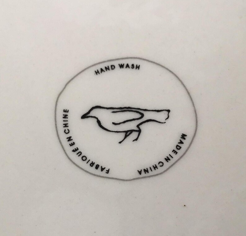 Puede incluir: Superficie de cer&aacute;mica blanca con un dise&ntilde;o gr&aacute;fico circular. El dise&ntilde;o presenta el contorno negro de un p&aacute;jaro y las palabras "HAND WASH", "FABRIQUE EN CHINE" y "MADE IN CHINA" en texto negro. El p&aacute;jaro mira hacia la derecha.