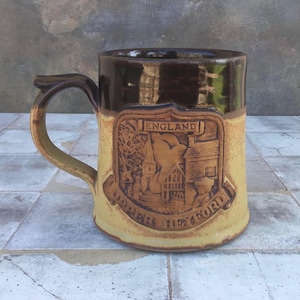 May include: A brown and tan ceramic mug with a raised design of a building and the text "ENGLAND" and "UPPER HEYFORD".