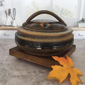 May include: A round, lidded ceramic casserole dish featuring a glossy, multi-toned glaze in shades of deep brown, amber, and muted blue. The lid includes a prominent arched handle, and the body has a tiered, circular design with a smooth finish.