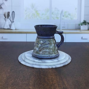May include: A handmade ceramic mug with a dark blue rim and handle, resting on a stack of three white marble coasters. The mug has a speckled, light green and brown body with blue accents. The background shows a kitchen setting.