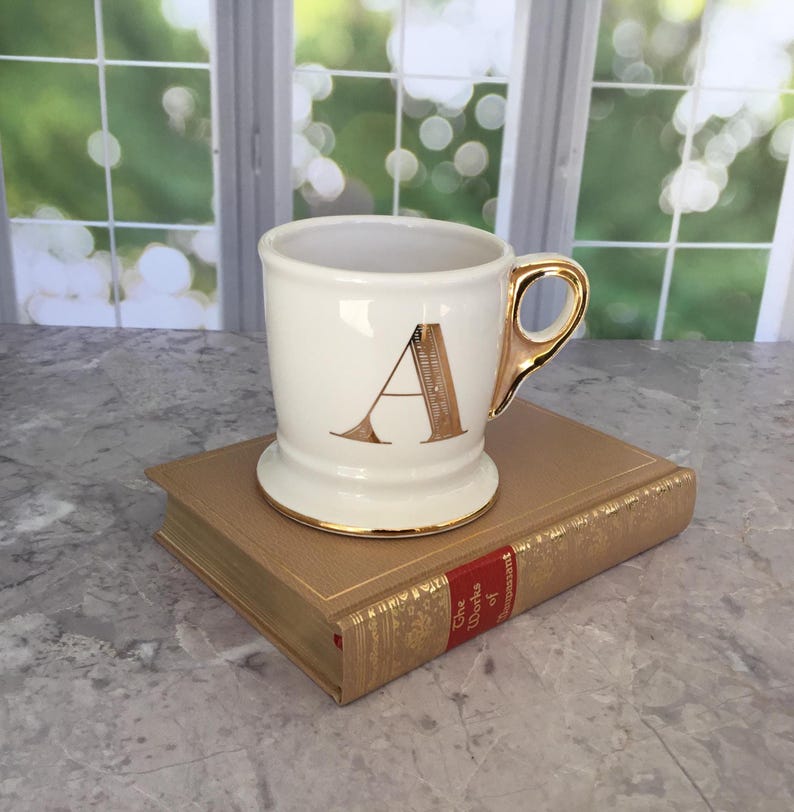 Puede incluir: Una taza de cer&aacute;mica blanca con un asa dorada y la letra "A" en escritura dorada. La taza est&aacute; sobre un libro dorado con letras rojas. La taza tiene un borde y una base dorados. El fondo incluye una ventana con una vista verde borrosa.