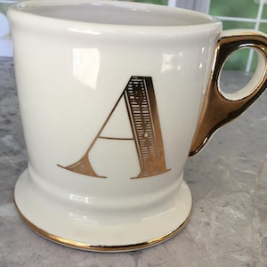 Puede incluir: Taza de cer&aacute;mica color crema con asa y ribetes dorados. La letra "A" dorada destaca en la parte delantera. La taza tiene una base de pedestal. Ideal para caf&eacute; o t&eacute;.