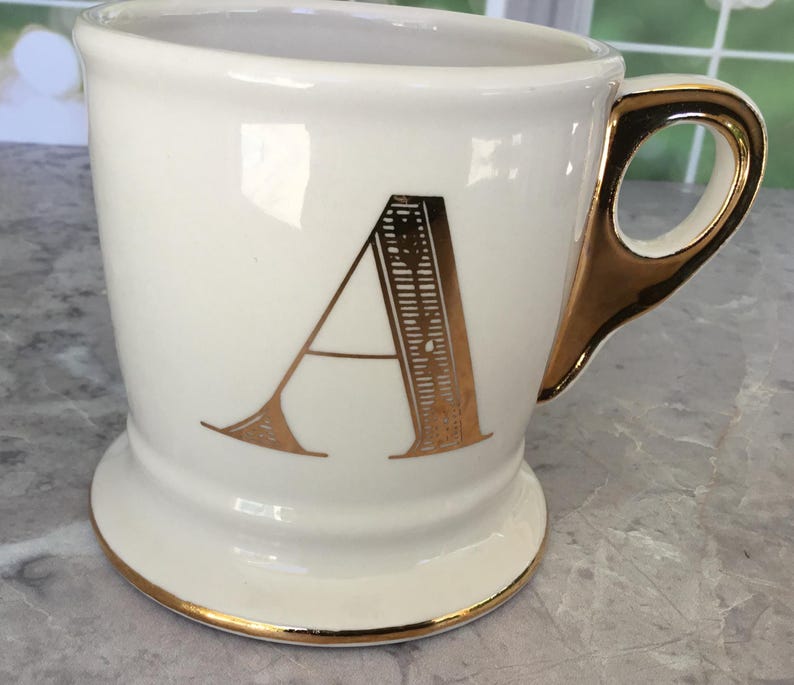 Puede incluir: Taza de cer&aacute;mica color crema con asa y ribetes dorados. La letra "A" dorada destaca en la parte delantera. La taza tiene una base de pedestal. Ideal para caf&eacute; o t&eacute;.
