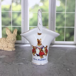 May include: White ceramic basket with a handle, featuring a Mrs. Rabbit illustration with her children. The basket has gold trim and the words "Mrs. Rabbit" printed on the front. A decorative bunny is in the background.