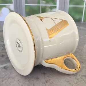 Puede incluir: Una taza de cer&aacute;mica blanca con borde y asa dorados. La taza presenta un dise&ntilde;o de la letra "A" dorada. La parte inferior de la taza tiene un logotipo en blanco y negro de un p&aacute;jaro. La taza est&aacute; sobre una superficie de m&aacute;rmol.