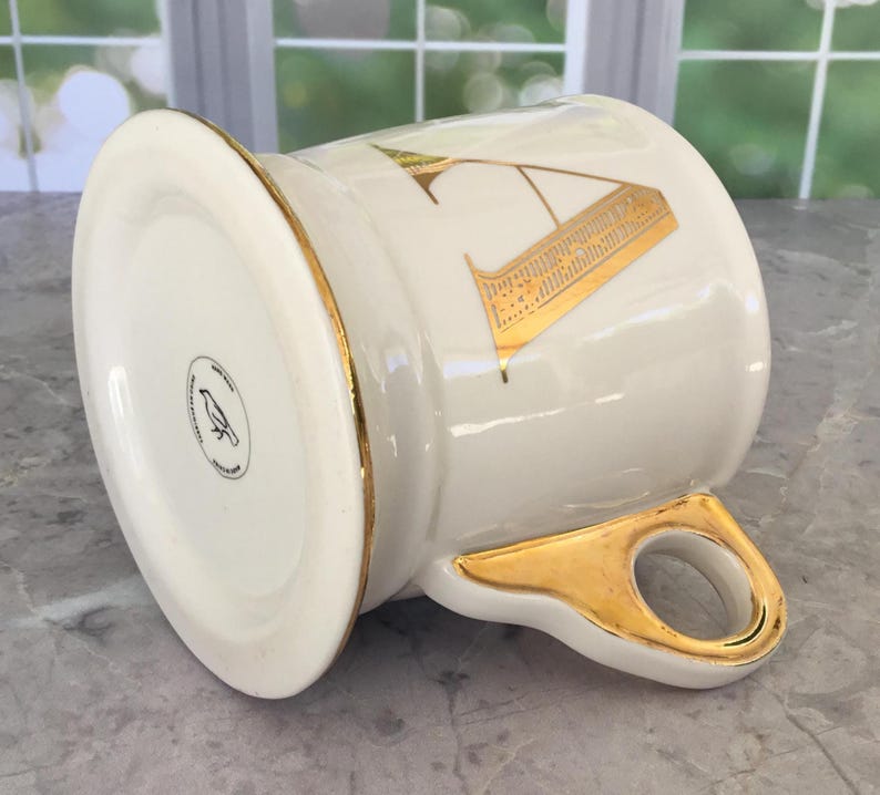 Puede incluir: Una taza de cer&aacute;mica blanca con borde y asa dorados. La taza presenta un dise&ntilde;o de la letra "A" dorada. La parte inferior de la taza tiene un logotipo en blanco y negro de un p&aacute;jaro. La taza est&aacute; sobre una superficie de m&aacute;rmol.