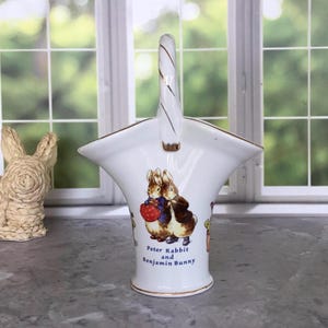 May include: A white ceramic basket with a handle, featuring a Peter Rabbit and Benjamin Bunny illustration. The basket has a gold trim and a curved handle with gold accents. The text "Peter Rabbit and Benjamin Bunny" is printed on the front.