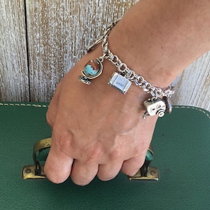 May include: A silver-toned metal charm bracelet featuring three distinct travel-themed charms: a small globe with blue and brown continents, a rectangular suitcase, and a vintage-style camper trailer, all attached to a classic link chain.