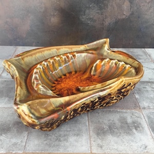 May include: A vintage ceramic ashtray with a unique, organic shape. The ashtray features a textured exterior in shades of brown and gold, with a vibrant orange and brown interior. The design is reminiscent of a seashell or a stylized leaf.