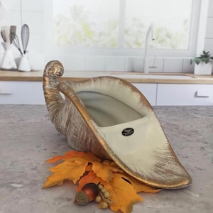 May include: A ceramic cornucopia vessel featuring a cream-colored interior and a textured exterior with a gold-toned finish. The piece includes a small oval black label with the text "Hagger" and is displayed with artificial orange maple leaves.