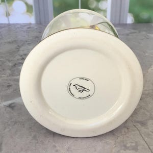 Puede incluir: Un jarr&oacute;n de cer&aacute;mica blanca con base redondeada y tapa de cristal transparente. La base del jarr&oacute;n tiene un emblema circular con una ilustraci&oacute;n de un p&aacute;jaro y las palabras "Hand Wash" y "Made in China".