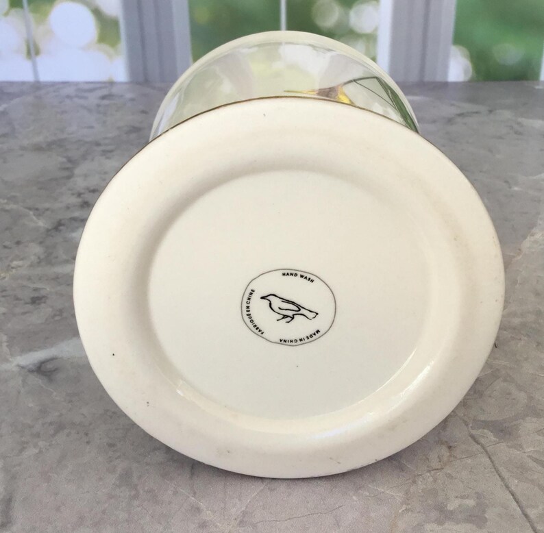 Puede incluir: Un jarr&oacute;n de cer&aacute;mica blanca con base redondeada y tapa de cristal transparente. La base del jarr&oacute;n tiene un emblema circular con una ilustraci&oacute;n de un p&aacute;jaro y las palabras "Hand Wash" y "Made in China".