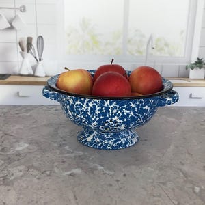 May include: A blue and white enamelware colander filled with several red and yellow apples. The colander has a pedestal base and two handles. The kitchen setting includes a window, countertop, and utensils.