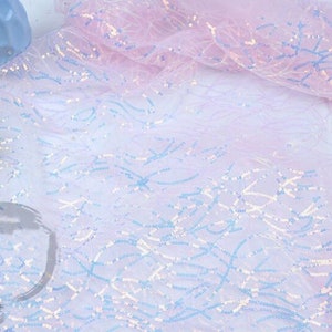 May include: A sheer, iridescent fabric with a light pink and white ombre effect. The fabric is decorated with small, blue and white sequins arranged in a wavy pattern.
