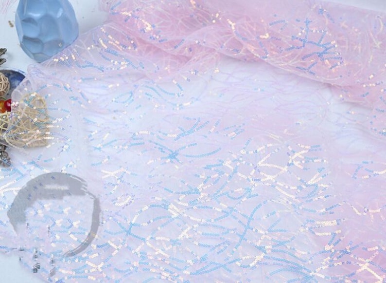 May include: A sheer, iridescent fabric with a light pink and white ombre effect. The fabric is decorated with small, blue and white sequins arranged in a wavy pattern.
