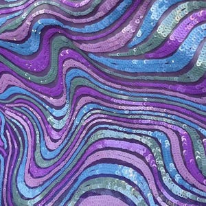 Può includere: Primo piano di un tessuto con un motivo ondulato di paillettes. Le paillettes sono in tonalità di viola, blu e verde, creando una superficie scintillante e strutturata. Il design ricorda l'acqua che scorre o un effetto psichedelico.
