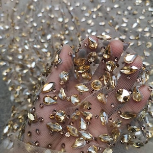 Può includere: Primo piano di un tessuto beige trasparente pesantemente decorato con strass dorati di varie dimensioni e forme. Gli strass sono sparsi sul tessuto, creando un effetto scintillante. Il tessuto è tenuto in mano, mostrando la trama e i dettagli.