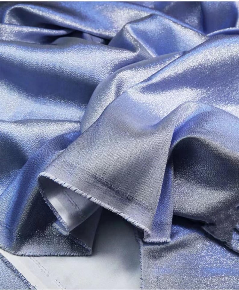 May include: Close-up of a light blue fabric with a subtle shimmer. The fabric has a smooth, silky texture and a slightly iridescent sheen.