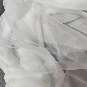 May include: Close-up of white tulle fabric, with a delicate, sheer texture. The fabric is draped and folded, creating shadows and highlights. The material appears soft and lightweight, suitable for various craft projects or apparel.