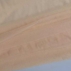 May include: Close-up of a folded, sheer fabric in a soft peach color. The fabric is layered, creating a textured effect with visible folds and subtle shadows. The material appears lightweight and delicate, suitable for various craft projects.