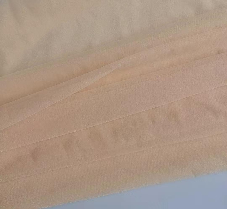 May include: Close-up of a folded, sheer fabric in a soft peach color. The fabric is layered, creating a textured effect with visible folds and subtle shadows. The material appears lightweight and delicate, suitable for various craft projects.