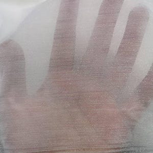 May include: A close-up of a sheer white fabric with a fine mesh texture. The fabric is slightly translucent, allowing a glimpse of the background. The material appears to be lightweight and delicate, with a subtle sheen.