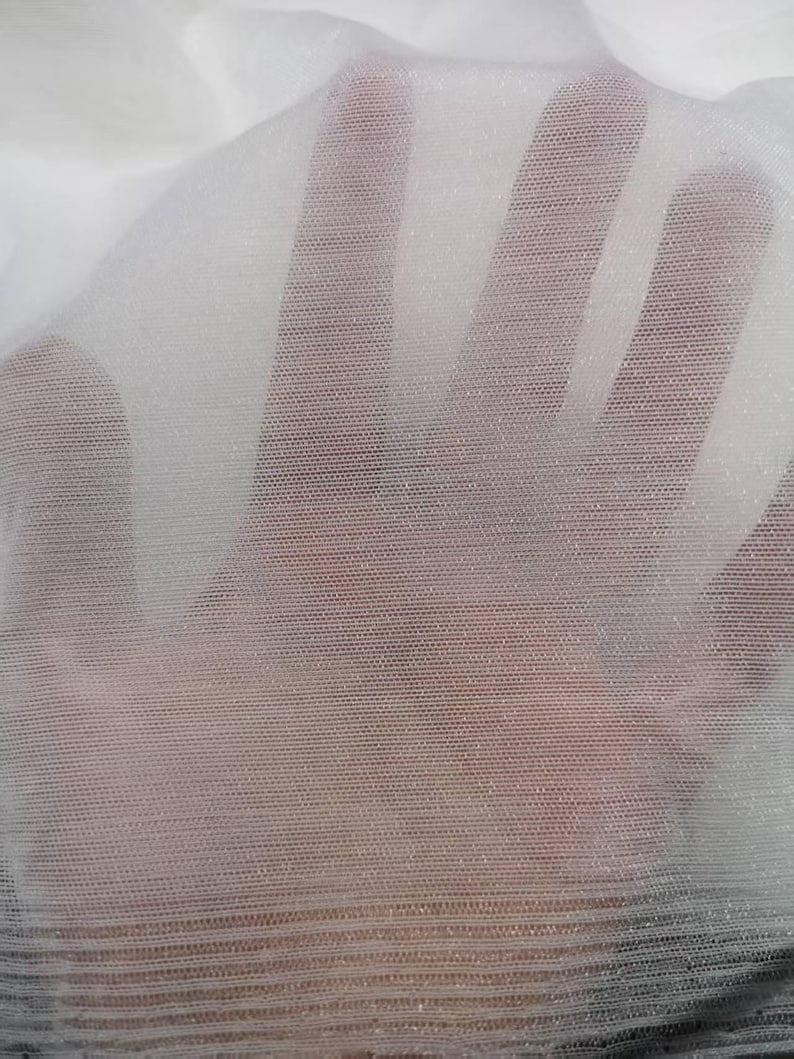 May include: A close-up of a sheer white fabric with a fine mesh texture. The fabric is slightly translucent, allowing a glimpse of the background. The material appears to be lightweight and delicate, with a subtle sheen.