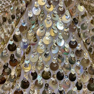 Pode incluir: Um tecido de malha branca densamente adornado com lantejoulas circulares douradas e prateadas. As lantejoulas estão dispostas em filas verticais, apresentando uma mistura de acabamentos metálicos reflexivos que criam uma textura brilhante e em camadas.