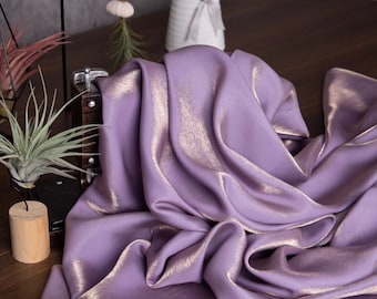May include: A draped, shimmering fabric in a soft lavender hue with gold highlights. The fabric is arranged in flowing folds, catching the light and creating a luxurious texture. The image suggests a textile for fashion or home decor.