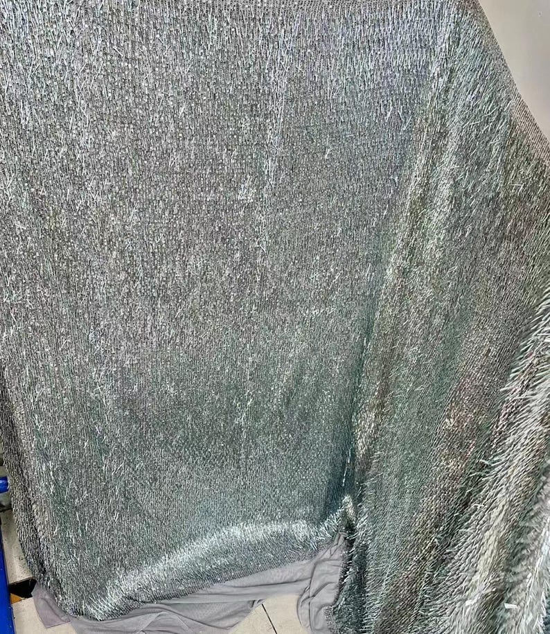 K&ouml;nnte beinhalten: Ein silberner, glitzernder Stoff mit einer strukturierten, flauschigen Oberfl&auml;che. Der Stoff ist in der Mitte gefaltet und zeigt die Innenseite und Au&szlig;enseite des Materials.